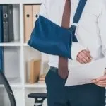 Cropped view of employee with broken arm in bandage standing and holding paper over table in office, workers' compensation concept.